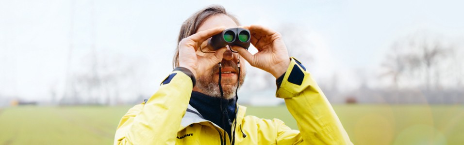 Das Bild zeigt einen Mann mit gelber Jacke, der durch ein Fernglas schaut.